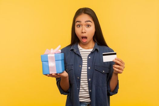 Wow gift shopping. Portrait of amazed girl in denim shirt holding present box, credit card and looking surprised, shocked by purchase, cashback and bank loan. studio shot isolated on yellow background