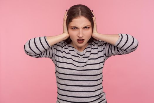 Don't want to hear you. Portrait of irritated woman in striped sweatshirt closing ears and looking angry, annoyed by loud sound, unpleasant noise. indoor studio shot isolated on pink background