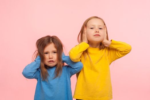 We don't want to listen. Portrait of two naughty disobedient little girls covering ears with hands, ignoring parental advice, avoiding annoying talk. indoor studio shot isolated on pink background