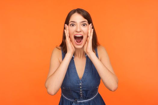 Oh my god, wow. Shocked impressed brunette woman in denim outfit standing with wide open mouth and clutching face in surprise, looking startled amazed at camera. indoor studio shot orange background