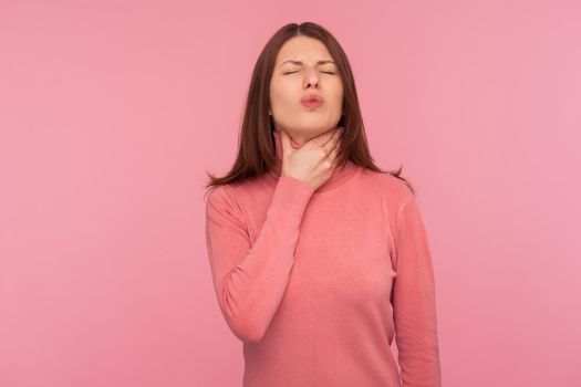 Sad woman with brown hair palping neck feeling throat ache and pain during swallow, tonsillitis inflammation. Indoor studio shot isolated on pink background