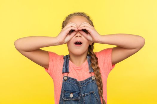 Amazed kid exploring world. Portrait of nosy little girl in denim overalls looking through fingers shaped like binoculars and expressing amazement, shocked by crazy event. indoor studio shot isolated