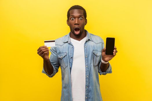 E-commerce. Portrait of surprised amazed man in denim casual shirt holding plastic bank card and phone, shocked by quick electronic money transfers. indoor studio shot isolated on yellow background