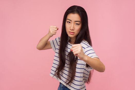 I'll punch you, Portrait of angry girl with long hair in striped t-shirt standing with boxing gesture, clenching fists ready to fight, threatening to attack. studio shot isolated on pink background