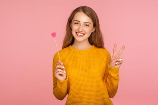 Portrait of joyful lovely ginger girl in sweater holding pink paper lips on stick, symbol of kiss and love, showing victory gesture and smiling at camera. studio shot isolated on pink background