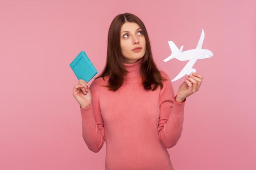 Thoughtful dreamy woman with brown hair holding passport and paper plane in hands planning her vacation abroad