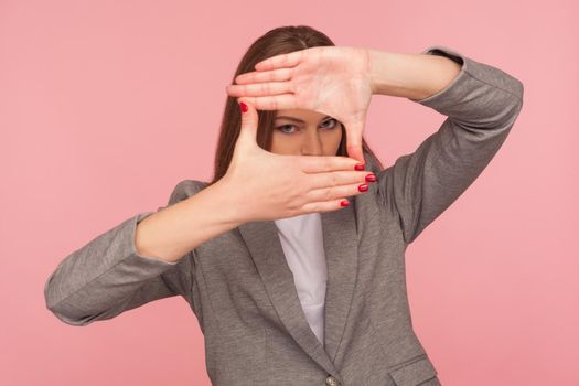 Portrait of purposeful young woman in business suit looking through finger frame, focusing on startup goal, capturing idea, watching perspectives. indoor studio shot isolated on pink background