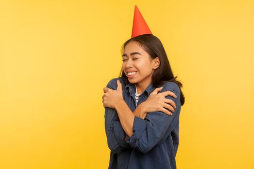 Happy birthday to me. Portrait of girl in denim shirt and with funny party cone on head embracing herself, celebrating successful event alone but happy. studio shot isolated on yellow background
