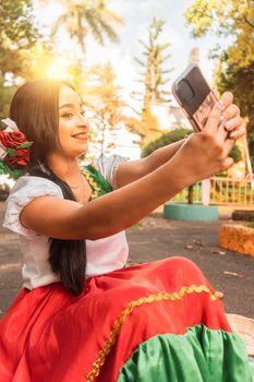 Medium shot of a beautiful Latin teenager in traditional dress taking a selfie in a park during a sunny day