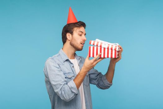 Portrait of curious man with party cone hat opening gift, peeking inside box with nosy look, unpacking present, in anticipation of interesting birthday surprise. studio shot isolated, blue background
