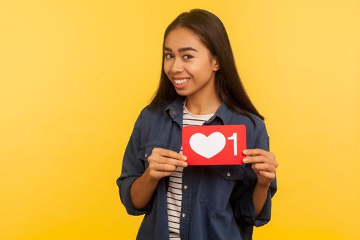 Portrait of playful girl in denim shirt showing heart Like icon, follower notification symbol, click button for social media, interesting content. indoor studio shot isolated on yellow background