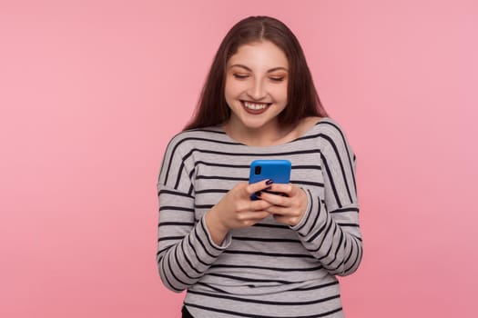 Portrait of happy smiling young woman typing on cell phone, reading funny blog, chatting in social network, using mobile app for online communication. indoor studio shot isolated on pink background