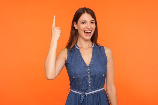 Smart brunette woman in denim dress pointing finger up with excellent idea, looking inspired and excited by sudden creative thought, found solution. indoor studio shot isolated on orange background