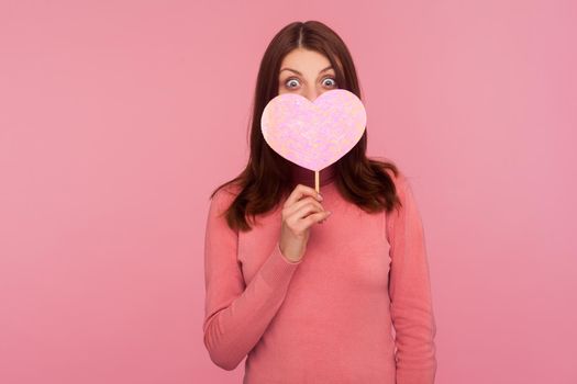 Surprised brunette woman in pink sweater covering face with pink heart, hiding behind symbol of love, wondered with romance date. Indoor studio shot isolated on pink background