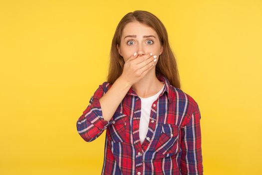 I won't tell anyone. Portrait of scared ginger girl in checkered shirt covering mouth with hand and looking frightened at camera, intimidated female afraid to talk. studio shot, yellow background