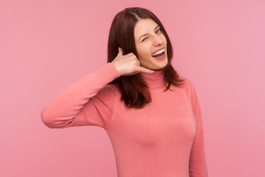 Cheerful playful woman with brown hair in pink sweater showing phone gesture and winking, asking to call her. Indoor studio shot isolated on pink background