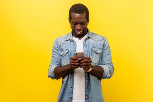 Portrait of happy smiley man in denim casual shirt using cellphone, typing text message or dialing number with joyous expression, technology concept. indoor studio shot isolated on yellow background