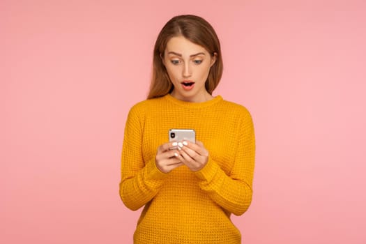 Portrait of surprised cute ginger girl texting on cellphone and expressing amazement, reading shocking message using smartphone, mobile application. indoor studio shot isolated on pink background