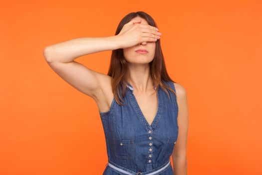 Don't want to look. Young brunette woman in denim dress covering eyes with hand, rejecting to watch forbidden inappropriate content, ignoring problems. indoor studio shot isolated on orange background