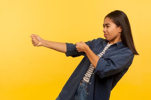 Portrait of diligent purposeful girl in denim shirt pulling invisible heavy burden, striving hard to achieve success, dragging with persistence. indoor studio shot isolated on yellow background