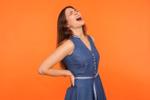 Terrible backache. Ill brunette woman holding lower back and screaming in pain, suffering injured spine disk with expression of acute unbearable ache. indoor studio shot isolated on orange background