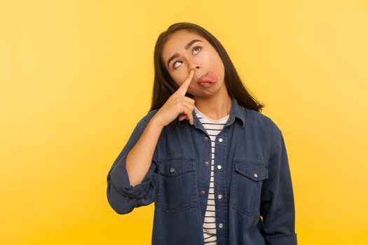 Bad habit, humorous grimace. Portrait of adorable funny girl in denim shirt picking nose and sticking out tongue with dumb comical stupid expression. indoor studio shot isolated on yellow background
