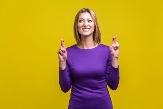 Portrait of worried beautiful woman in elegant purple dress holding fingers crossed for good luck, hoping for fortune, longing for wishes come true. indoor studio shot isolated on yellow background
