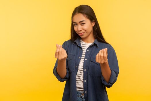 Portrait of greedy girl in denim shirt showing money gesture and looking with cunning smile, planning big profit, financial reward, demanding cash. indoor studio shot isolated on yellow background