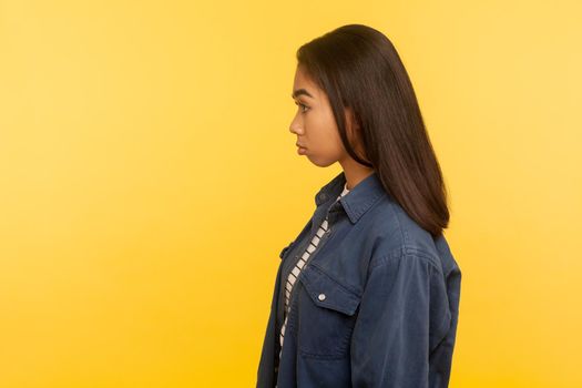 Side view of upset girl in denim shirt standing with unhappy gloomy expression, feeling annoyed and dissatisfied with defeat, negative emotions. indoor studio shot isolated on yellow background