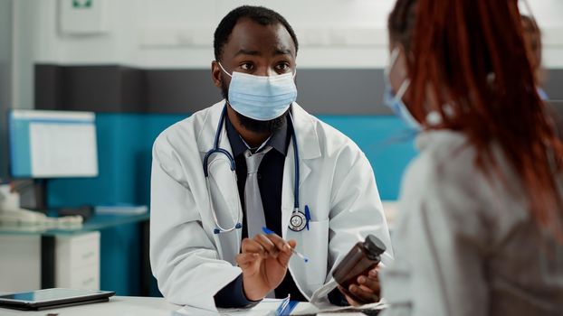 Maternity specialist and pregnant woman with face mask at checkup visit, meeting to talk about pregnancy. Future mother with baby bump asking for medical advice from physician at cabinet desk.