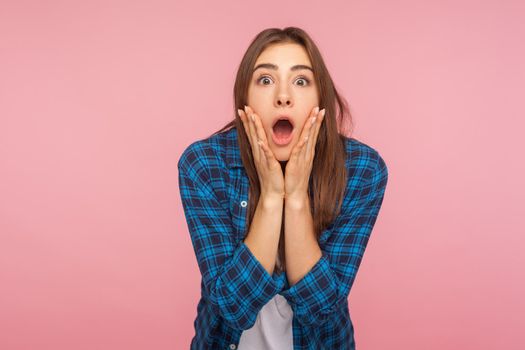 Portrait of astonished emotional funny girl in checkered shirt looking with big eyes, holding hands on face and screaming in amazement, shocked by sudden event. studio shot isolated on pink background