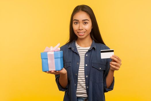 Gift shopping. Portrait of happy girl in denim shirt holding present box, credit card and smiling to camera, satisfied with purchase, cashback and bank loan. studio shot isolated on yellow background