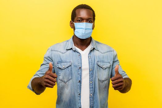 Thumbs up, excellent job. Portrait of enthusiastic handsome man in denim casual shirt with surgical medical mask smiling and showing like gesture at camera. studio shot isolated on yellow background