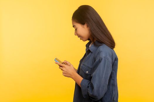 Side view of happy girl in stylish denim shirt typing message and smiling, dialing number calling on cell phone, using online mobile application, searching web. indoor studio shot, yellow background