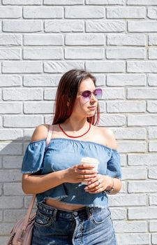 Summer vacation, street food eating. Charming stylish woman, enjoying drinking coffee outdoors, dressed in jeans shirt, wearing sunglasses on white brick wall background