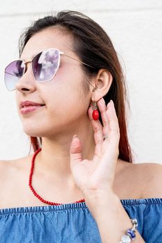 stylish young woman wearing jeans shirt, sunglasses and red jewelry at street, white wall background, summer look, copy space