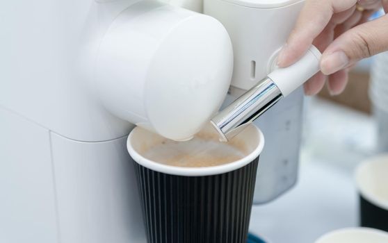 Woman making a cup of hot coffee with capsule coffee machine. Woman hand holding frothed milk dispenser of capsule coffee machine on table. Espresso coffee maker. Morning drink. Modern home equipment.