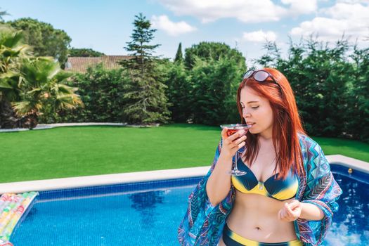 young red-haired woman drinking cherry cocktail standing by a swimming pool, wearing sunglasses, bikini and sarong. attractive girl enjoying her summer holiday. leisure and travel concept. natural sunlight, outdoor.