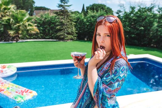 attractive young woman drinking a soft drink standing by the pool looking at camera. girl on her summer holiday, wearing sunglasses, bikini and sarong. leisure and travel concept. natural sunlight, outdoor.