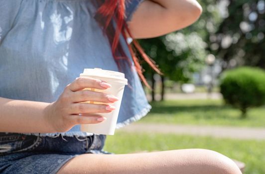 Attractive young woman in summer clothes and sunglasses holding cup of coffee in her hands while sitting on bench in the park