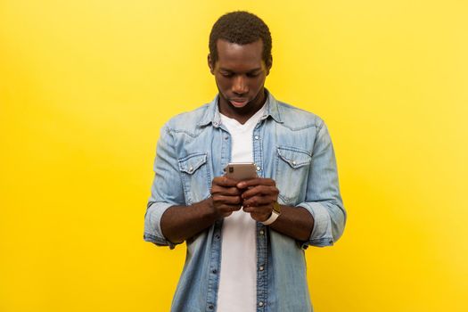 Portrait of serious man in denim casual shirt using cellphone, typing text message or dialing number with attentive expression, technology concept. indoor studio shot isolated on yellow background