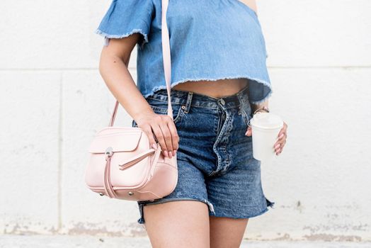 closeup of stylish woman with summer look, wearing jeans shorts, pink bag and holding cup of coffee, copy space