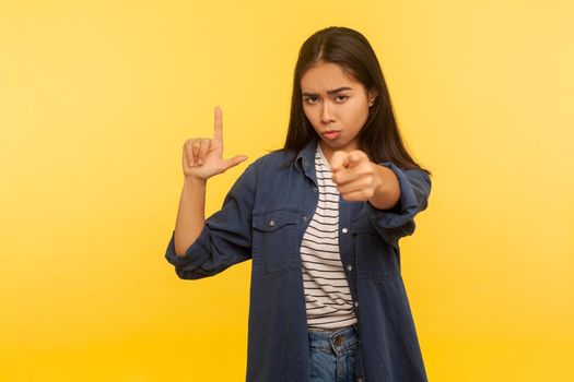 Hey you loser. Portrait of girl in denim shirt making, L sign and pointing to camera saying you lost, accusing for failure unsuccess, firing from job. indoor studio shot isolated on yellow background