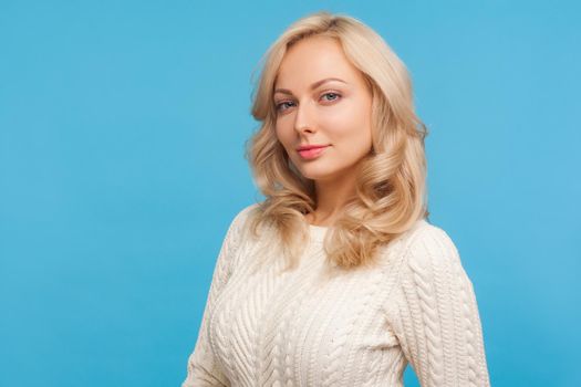 Portrait of sensual free woman with curly blond hair in white knitted sweater seriously looking at camera, femininity. Indoor studio shot isolated on blue background
