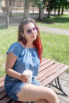 Attractive young woman in summer clothes and sunglasses holding cup of coffee in her hands while sitting on bench in the park