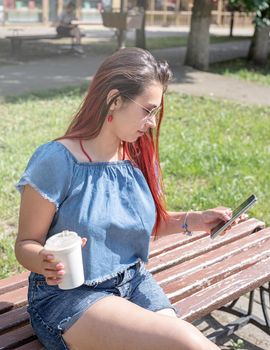 Attractive young woman in summer clothes and sunglasses holding cup of coffee in her hands,using smartphone videocall while sitting on bench in the park