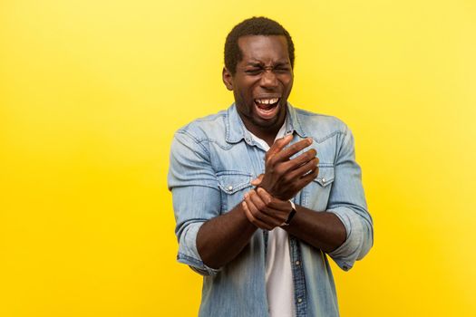 Portrait of sick or injured man in denim shirt standing with closed eyes, holding hand and shouting from pain in sprained wrist, suffering arthritis. indoor studio shot isolated on yellow background