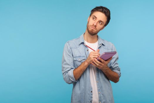 Portrait of pensive man journalist in denim shirt standing with thoughtful expression and holding paper notebook, pondering business idea, future plans. indoor studio shot isolated on blue background