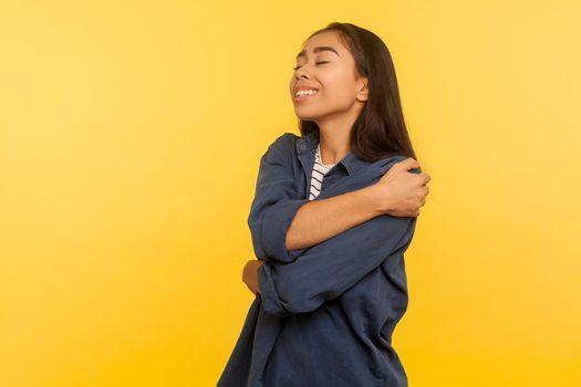 I love myself. Portrait of satisfied egoistic happy girl in denim shirt embracing and supporting herself, closed eyes with pleasure, being selfish, narcissistic. indoor studio shot, yellow background