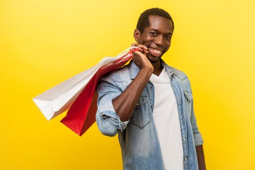 Portrait of satisfied buyer, positive man with attractive toothy smile in denim casual shirt holding shopping bags on his shoulder, enjoying discounts. indoor studio shot isolated on yellow background